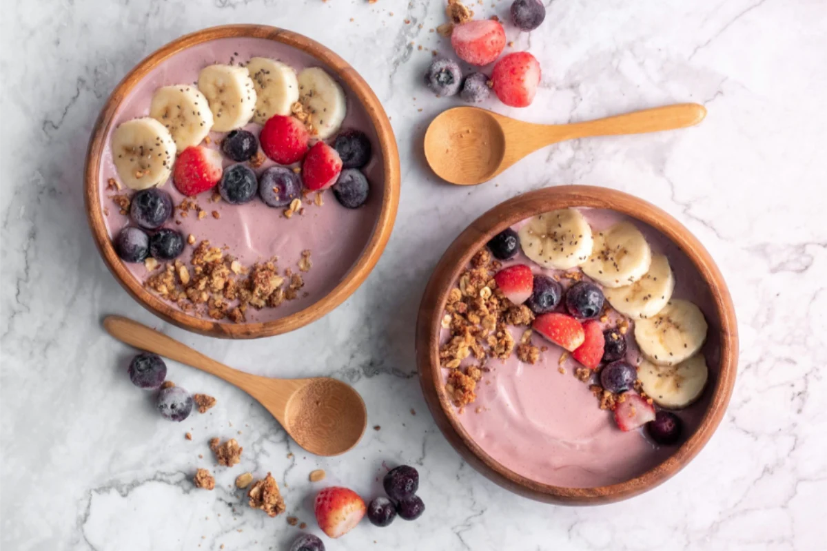 Eine braune Holzschüssel mit pflanzlichem Erdbeer-Joghurt, Banane, Beeren, HAFERVOLL Flapjack Cranberry & Cocoa Nib, Chiasamen und Agavendicksaft.