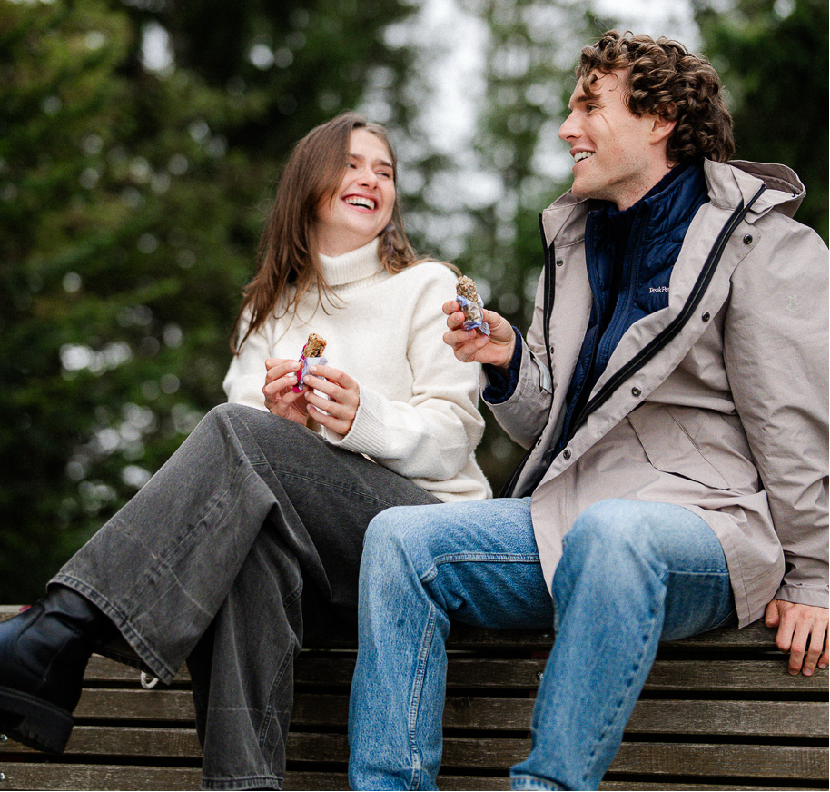 Ein Mann und eine Frau sitzen auf einer Bank und lachen sich an. Beide halten einen HAFERVOLL Flapjack in der Hand.